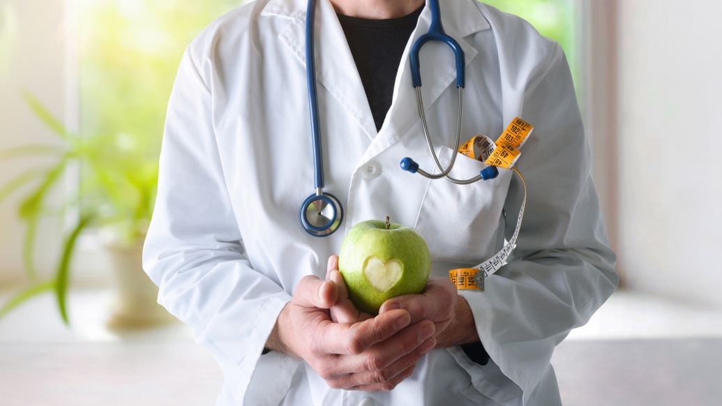 apple in hand of doctor with white coat and stethoscope facing frontward, neck down, and green plant with sunlight behind it from window coming through on white windows sill