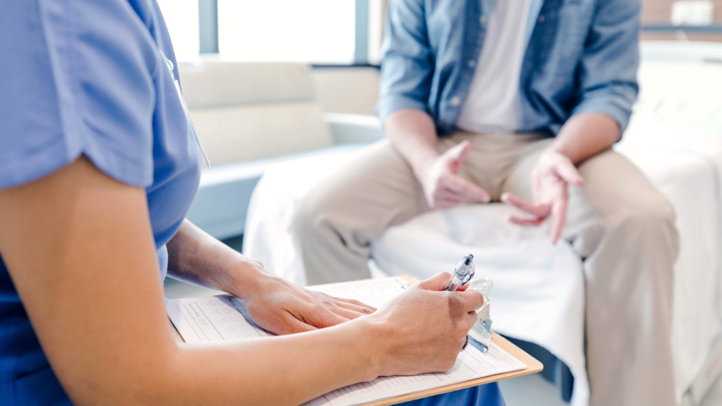 provider sitting writing something down with person in khakis and blue shirt unbuttoned with white shirt under