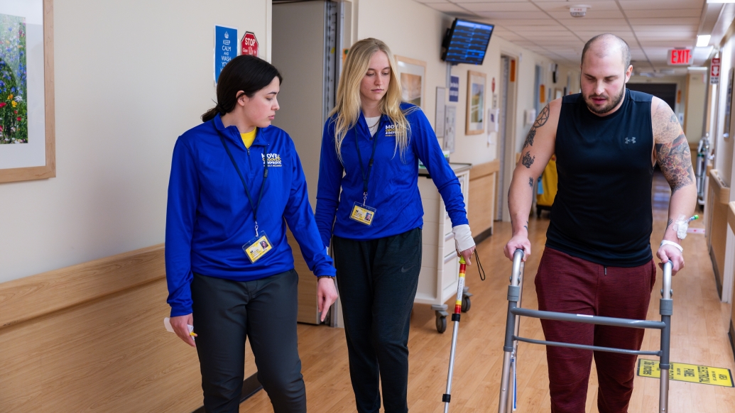 two students in bright blue jackets walking with man with walker down hallway in black sleeveless shirt and maroon sweats