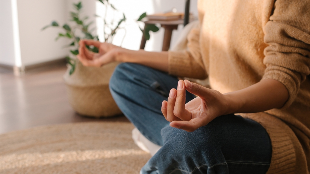 meditating person hands up on knees in jeans and camel sweatshirt on brown tan rug with plant in background and white wall