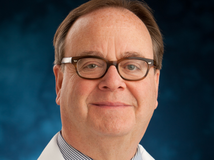 Middle aged white man with brown hair and glasses, wearing a business shirt, tie, and white doctor's coat with his name (Alan Dengiz) on it