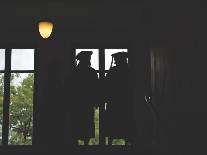 This is a dark photo of two graduates wearing graduation gowns, and caps with tassels. They are silhouetted in front of a window. 