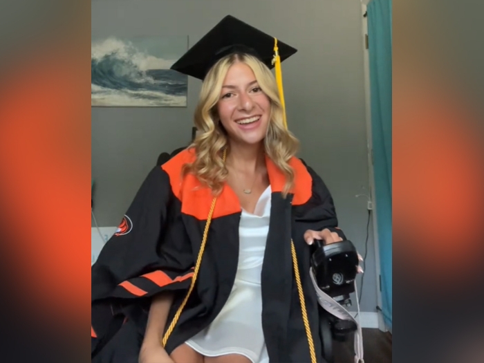 student sitting in white dress, cap and gown black with orange sitting