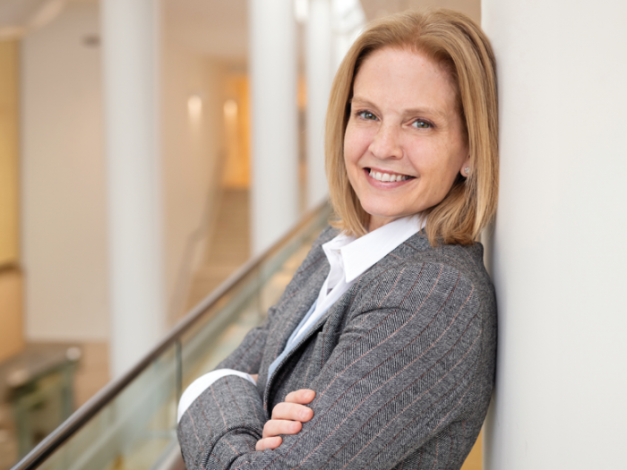 Headshot of Carrie Cunningham. She's wearing a gray suit jacket with a white shirt. She has shoulder-length blond hair and is leaning against a wall with her arms crossed. 