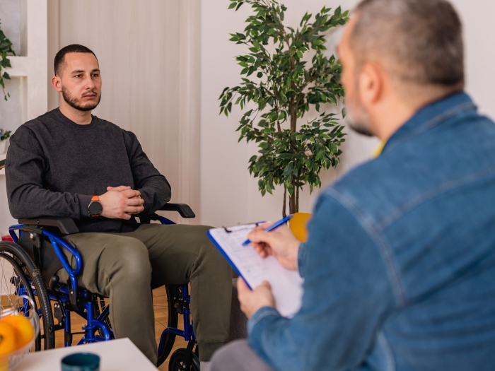 man in wheelchair talking to other man with back to camera in jean shirt in living room looking setting with tree in corner