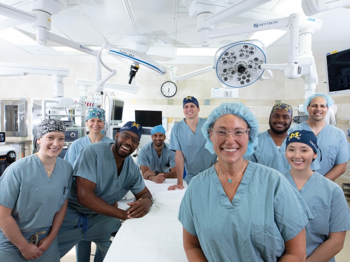 Michelle Caird, M.D., stands in an operating room with eight faculty and residents. They are all smiling and wear blue scrubs. They surround an empty operating table. 