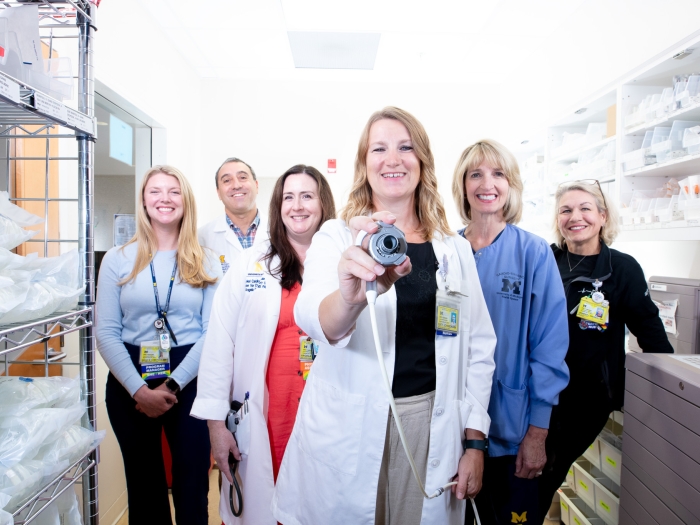 Caty Johnson, RN, DNP, holds a left ventricular assist device in her right hand, extending it towards the viewer. Five smiling clinicians stand behind her.