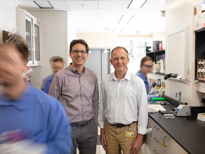 Scott Soleimanpour, M.D., and Lonnie Shea, Ph.D., stand in a lab smiling and facing the viewer. Three lab members work around them, appearing blurred and in motion.