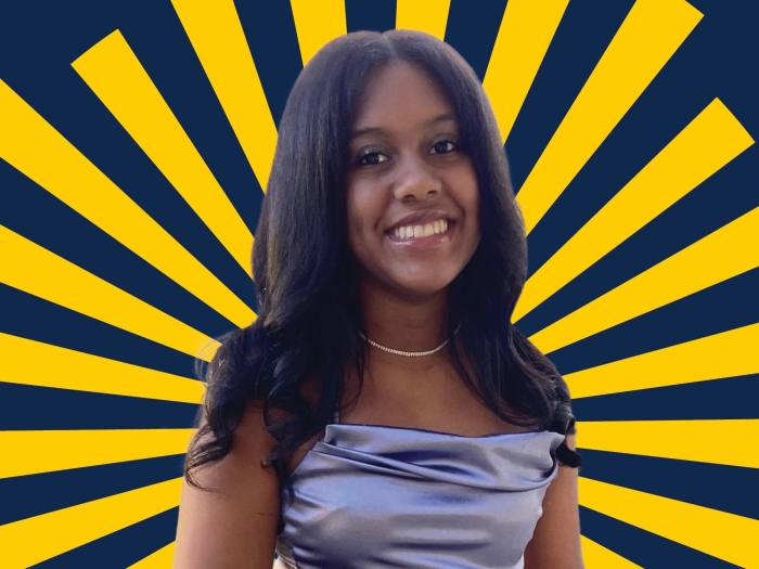 Portrait of Madison smiling and surrounded by gold rays. She has long black hair parted down the middle and is wearing a simple necklace and a formal silver top. 