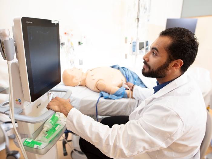 Medical student George Yacoub sits in front of a monitor in the Clinical Simulation Center. A medical training mannequin is on a bed in the background.