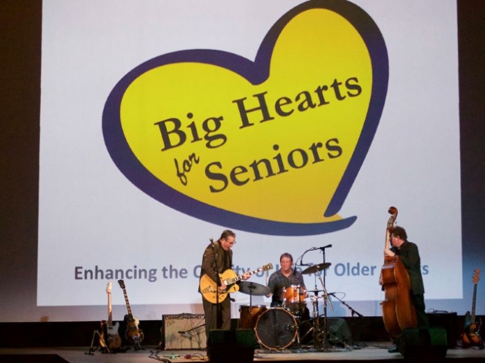 A musical trio dressed in black performs on a stage. One plays an electric guitar, one plays a drum set, and the third plays upright base. Three electric guitars on stands surround them. A large yellow heart with the text "Big Hearts for Seniors" is projected on the wall behind them. 