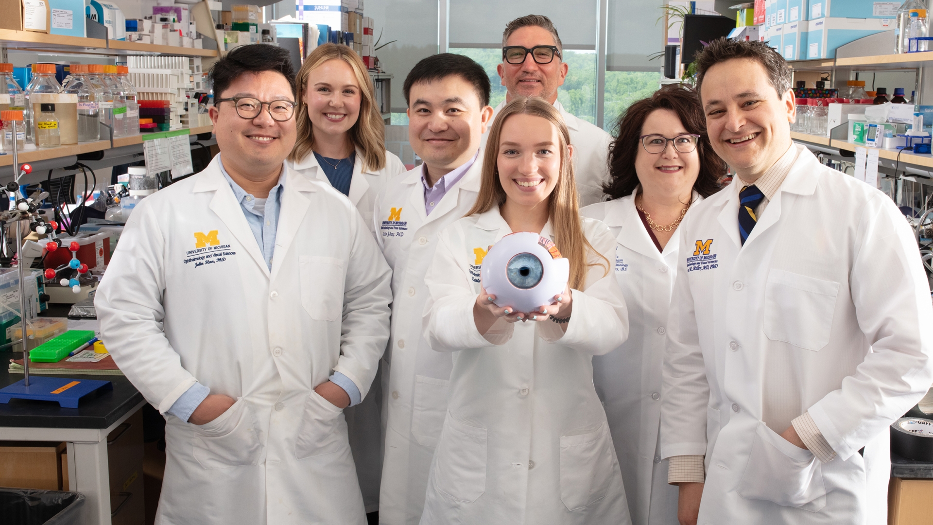 Jason Miller, M.D., right, and six members of his lab team smiling, wearing white coats, and standing in a lab. A woman in the middle extends a model of an eyeball towards the viewer.