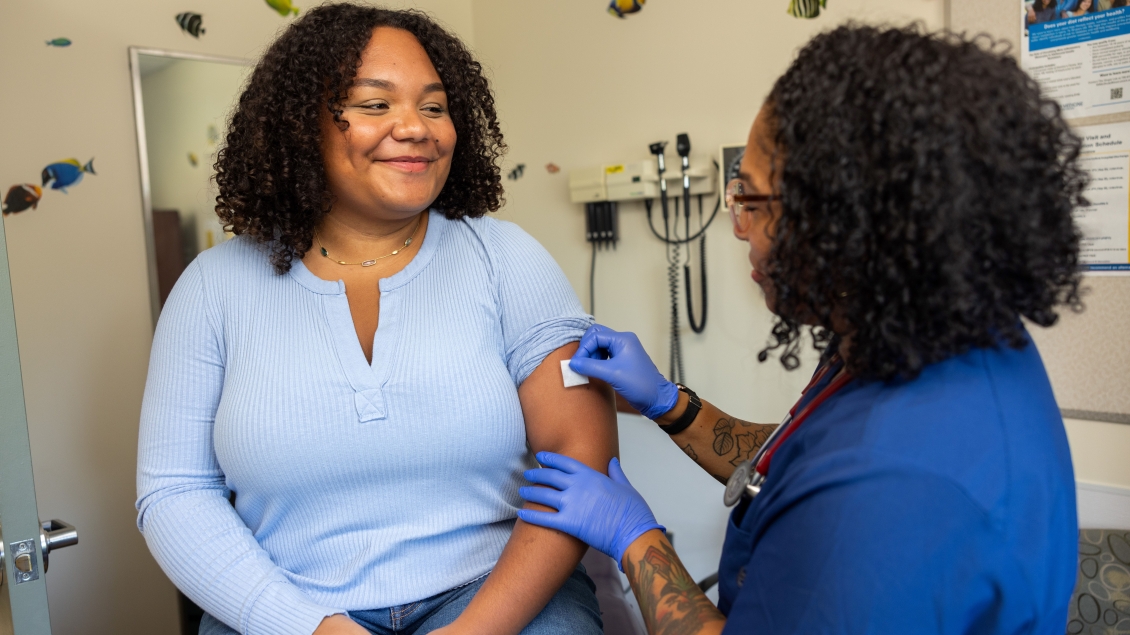 A young Black woman smiles as she is prepared for an injection by a tattooed multiracial able-bodied woman provider.