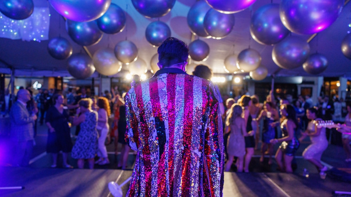 View from behind of a person in a red and white sequin-striped jacket on a stage at a party. Silver spheres hang from the ceiling. A large group of people in semi-formal attire stand facing in various directions, some walking and some talking in groups.