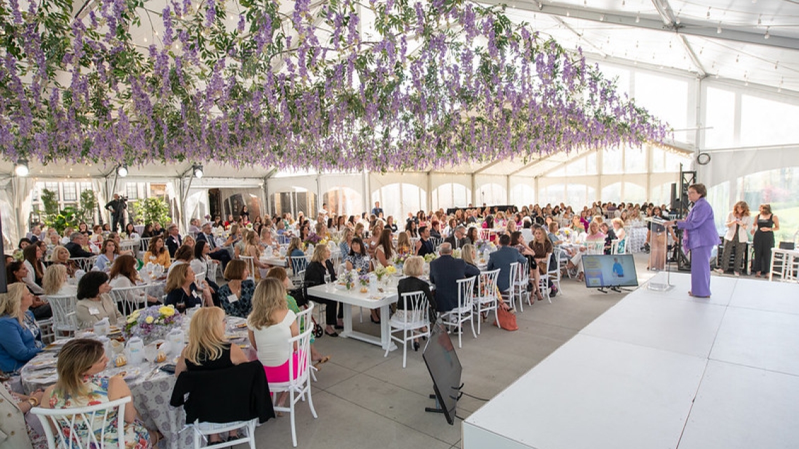 A large group of Women's Health Luncheon attendees seated around tables in a spacious, light-filled room.  They are listening to a speaker who is presenting from an elevated platform.