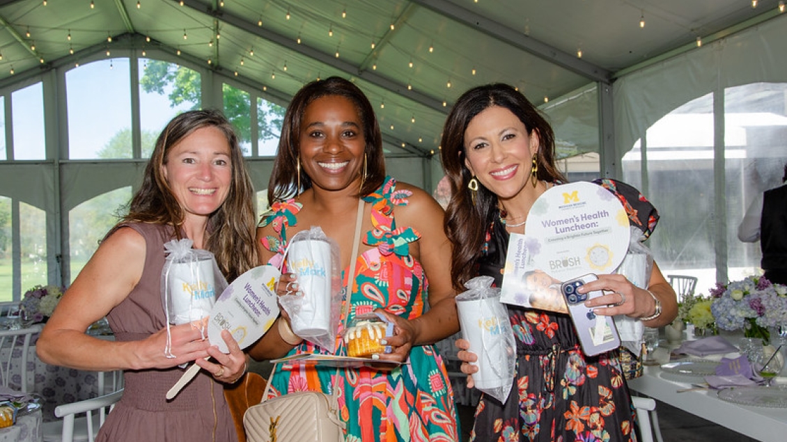 Three smiling Women's Health Luncheon attendees holding U-M gear, including coffee cups and hand-held fans.