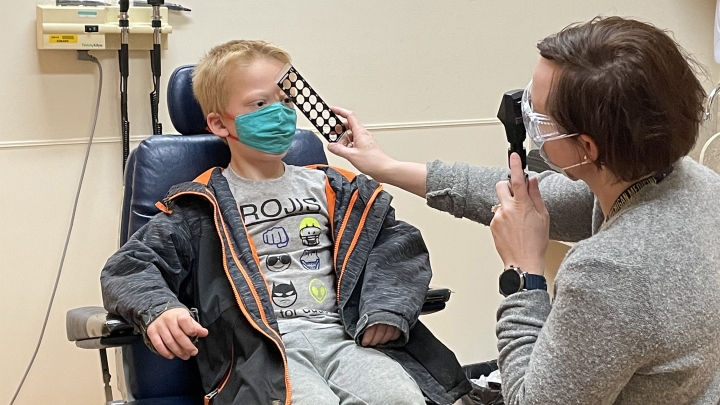 Young blond-haired boy seated in chair wearing jacket and mask getting eye exam