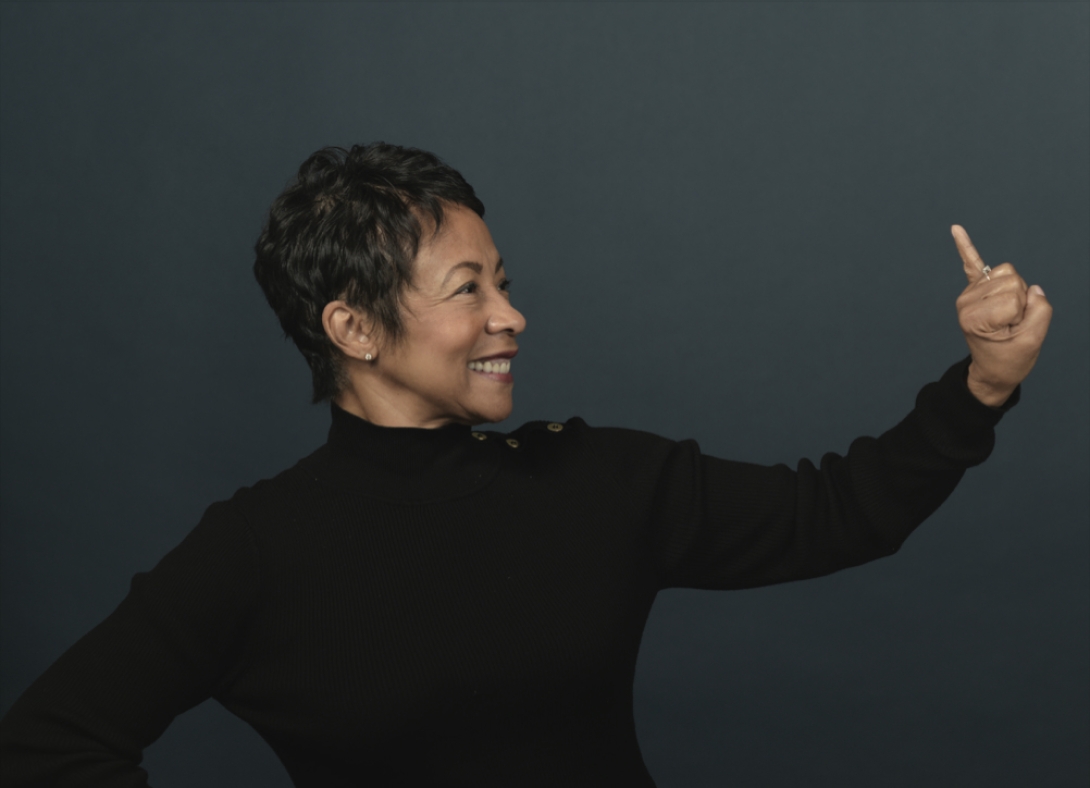 Portrait of Felicia McDaniel. She's wearing a black turtleneck sweater and standing in front of a dark gray background. She's facing to the side and is raising her left pinky in a sorority hand sign.