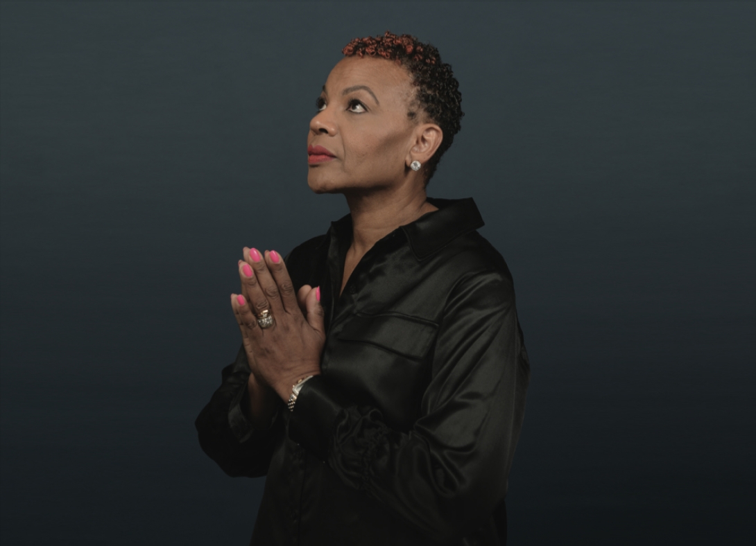 Portrait of Tamara Myles. She is wearing a black shirt and it standing in front of a dark gray backdrop. Her head is facing upwards and her hands are held in a prayer.