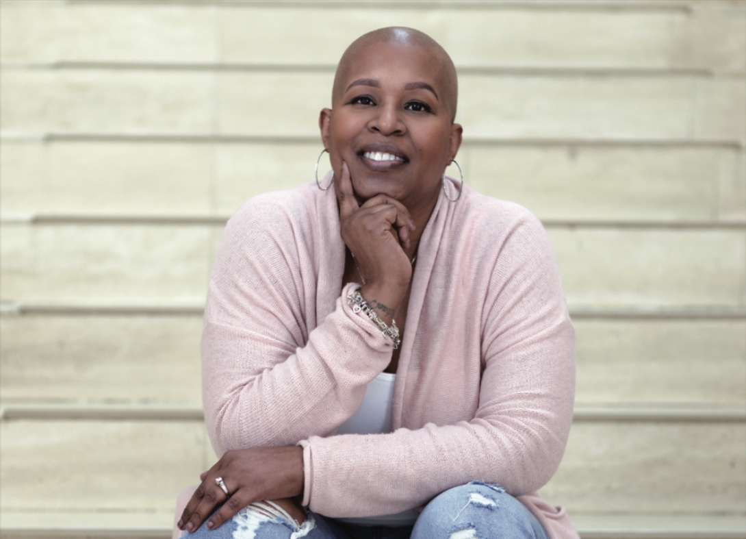 Portrait of Tanisha Kennedy. She's sitting on tan marble stairs and has her left arm resting on her legs. Her head is propped up by her hand underneath her chin.