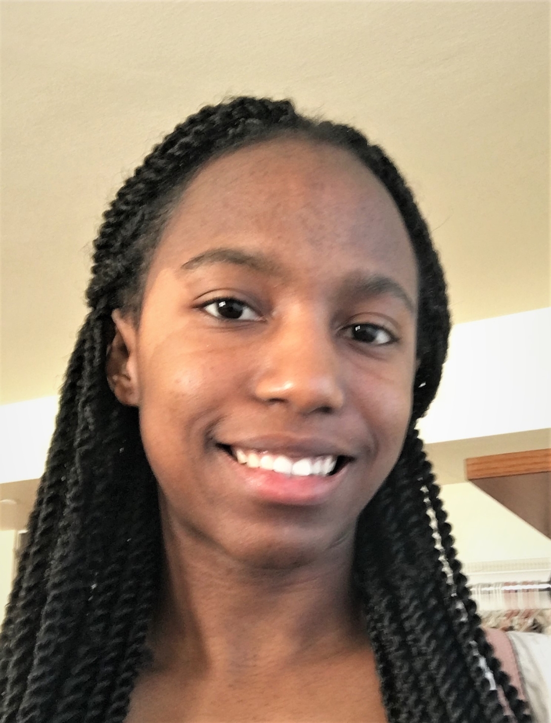 Laura describers herself as having black skin, brown eyes, mid-length black hair, and a large forehead. She's smiling in front of a white wall.