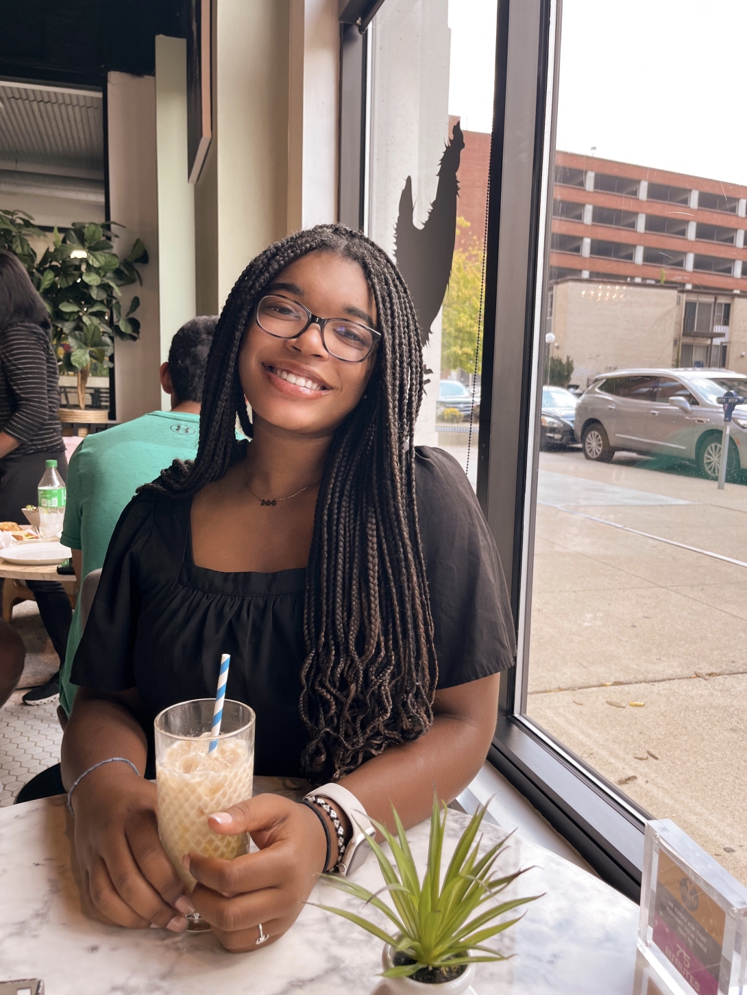 Miracle describes herself as brown skin, brown eyes, black hair, and glasses. She's wearing a black top and smiling at a table in a cafe with a drink in front of her.