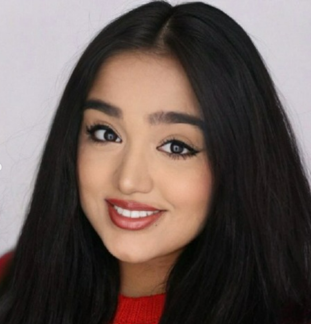 Samreen describes herself as having light brown skin, dark brown eyes, and black hair. She's wearing a red top and smiling in front of a grey background.