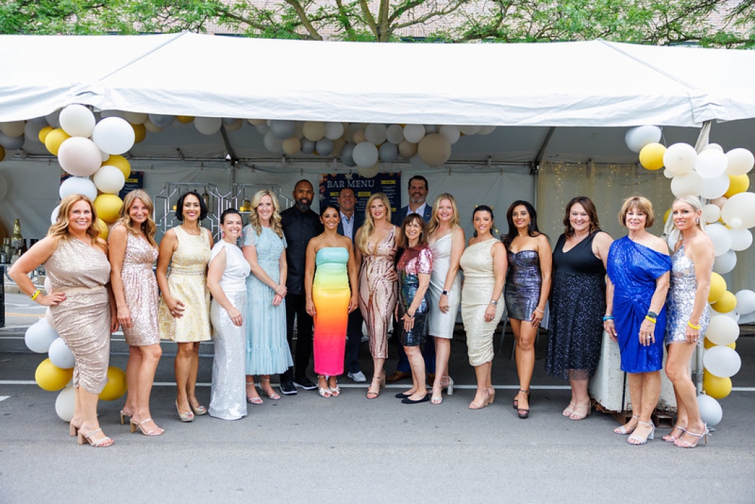 Seventeen smiling people in semi-formal attire stand in a semicircle under a white, open-sided party tent outside. Two columns of white, silver, and gold balloons are at each end of the line, with more balloons visible under the tent ceiling.