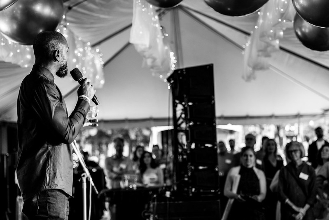 A black and white image of an outdoor party under a tent. On the left, Charles Woodson speaks into a microphone to a large group of standing guests. Hanging from the ceiling are shiny decorative spheres and ruffled drapes with decorative lights.