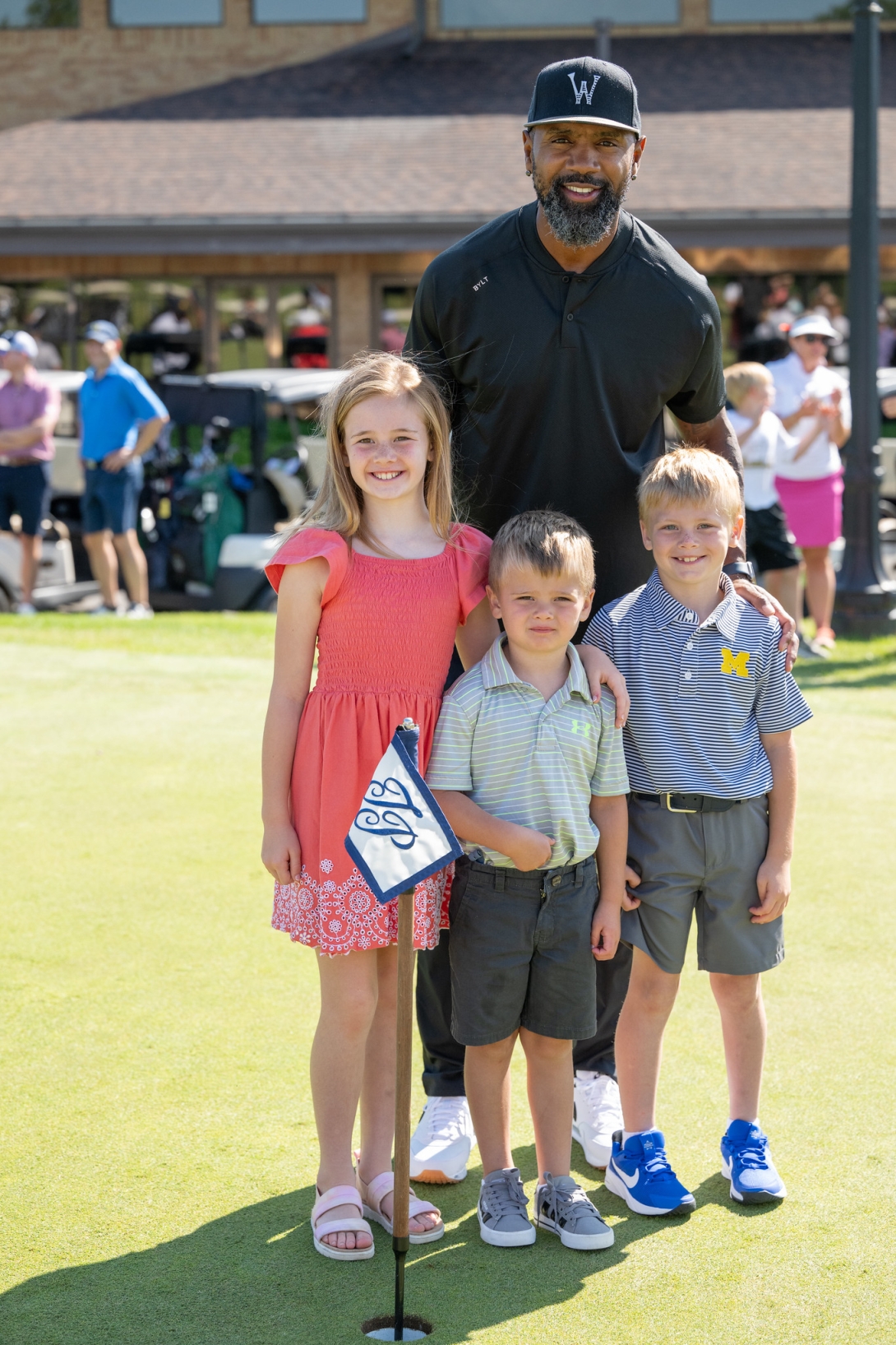 Charles Woodson stands behind three smiling children on a sunny golf course. A golf hole with a blue and white flag with the letters "T" and "P" is in front of them. Golf carts, people in summer attire, and a brown building with large windows are in the background.