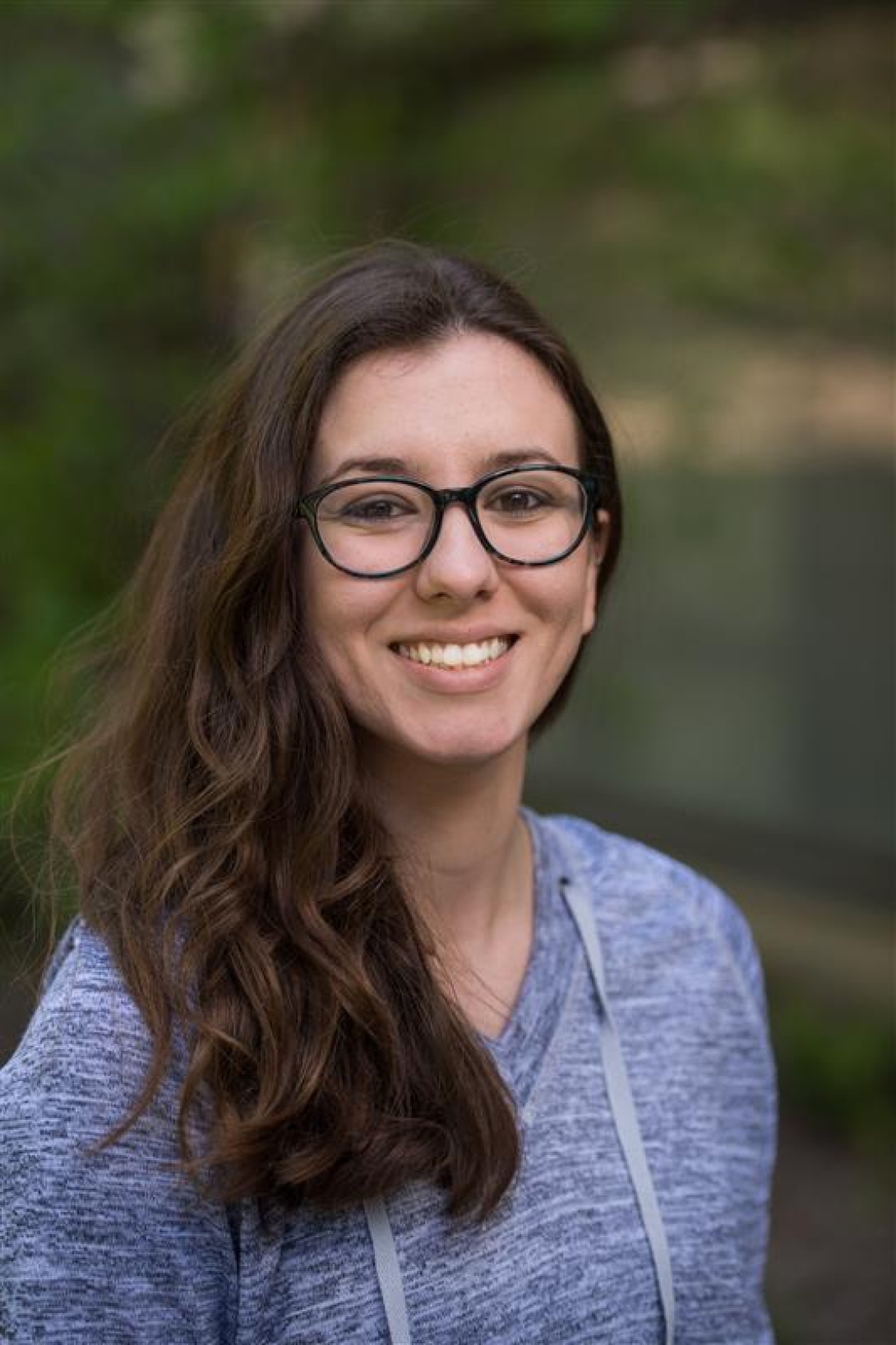 A photo of Isabelle Birt smiling in front of a natural background. Isabelle describes herself as having pale skin, brown eyes, dark brown hair, and wide-framed glasses.